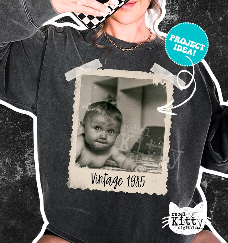 May include: Dark gray sweatshirt featuring a vintage photo print of a baby with the text "Vintage 1985." The photo is taped to the sweatshirt with white tape. A teal circle with the words "PROJECT IDEA!" is also on the sweatshirt.