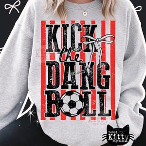 May include: A light gray sweatshirt with the text "KICK the DANG BALL" in distressed black lettering. The design includes a soccer ball and red vertical stripes. A white bow is also part of the design.