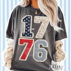 Könnte beinhalten: Dunkelgraues T-Shirt mit den aufgenähten Zahlen "1776" auf der Vorderseite. Die Zahlen bestehen aus verschiedenen patriotischen Mustern, darunter Sterne und Streifen, Blumenmuster und Vichy-Karo. Das Shirt hat eine lockere Passform und kurze Ärmel.