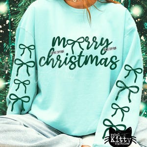 May include: A light blue sweatshirt with the words "Merry Christmas" in dark green script. The sleeves are adorned with dark green bow graphics. The bottom right corner features a black cat logo with the text "rebel kitty digitals".