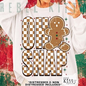 May include: A white long-sleeve sweatshirt featuring a large graphic with the text OH SNAP in a brown and white checkered pattern. A brown gingerbread person with a broken leg and three small buttons is positioned next to the text.