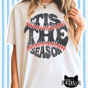 May include: Cream-colored t-shirt with a distressed baseball graphic. The graphic reads "'TIS THE SEASON" in a retro font, with a baseball design in the center. The shirt has a relaxed fit and a small black cat logo at the bottom.
