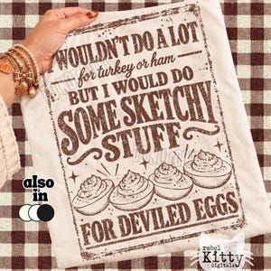 May include: A cream-colored t-shirt featuring a brown distressed graphic with the text: WOULDN'T DO A LOT for turkey or ham — BUT I WOULD DO SOME SKETCHY STUFF FOR DEVILED EGGS. The design includes an illustration of four deviled eggs.