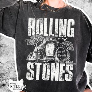May include: Dark gray sweatshirt with the words "ROLLING STONES" in a distressed white font. The graphic includes a cross, tomb, trees, and the text "He is Risen! Since AD 33". The sweatshirt has a vintage, worn look.