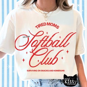 May include: Cream-colored t-shirt with the words "Tired Moms Softball Club" in red script. The shirt also features the phrase "Surviving on Snacks and Homeruns" and a black cat logo. The shirt is a casual wear item.