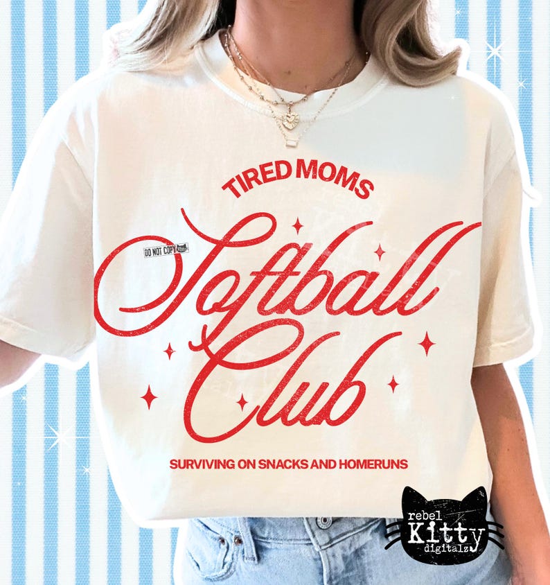 May include: Cream-colored t-shirt with the words "Tired Moms Softball Club" in red script. The shirt also features the phrase "Surviving on Snacks and Homeruns" and a black cat logo. The shirt is a casual wear item.