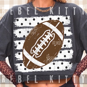 May include: A charcoal gray crewneck sweatshirt featuring a large brown football graphic centered over a white rectangular background with black polka dots. The design includes white distressed stripes on the football and the text REBEL KITTY.