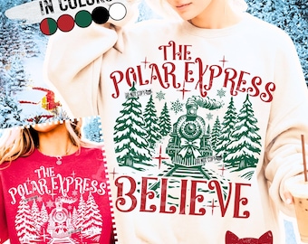 May include: Cream-colored sweatshirt and red t-shirt featuring a graphic of a train, pine trees, and the words "The Polar Express" and "Believe." The design includes snowflakes and the text "Do Not Copy."