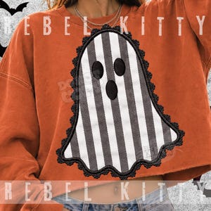 Peut inclure: Un sweat-shirt orange à manches longues avec une grande applique en forme de fantôme à rayures verticales noires et blanches. Le fantôme a trois yeux et une bouche ovales noirs, bordés d'une dentelle festonnée noire. Le texte REBEL KITTY est visible.