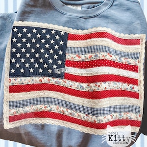Peut inclure: Un sweat-shirt bleu clair orné d'un drapeau américain en patchwork. Le drapeau présente un champ bleu denim avec des étoiles blanches, des rayures rouges et blanches et une bordure en dentelle. Le texte "DO NOT COPY" est visible.