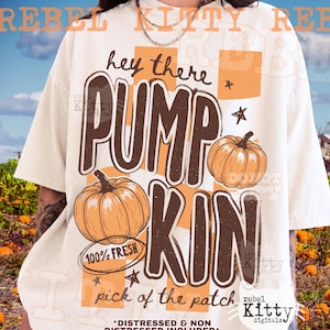 May include: An off-white t-shirt featuring a graphic with the text "hey there PUMP KIN" in brown, "100% FRESH" in a circle, and "pick of the patch" at the bottom. The design includes two orange pumpkins and orange checkered squares.