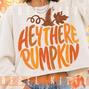 May include: A cream-colored cropped sweatshirt featuring a graphic print of a brown pumpkin stem and leaf above the orange and yellow text "HEY THERE PUMPKIN" in a brushstroke font. The garment has long sleeves and a crew neckline.