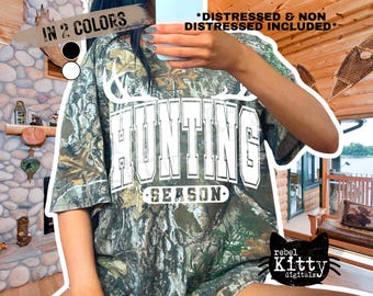 May include: Camouflage-patterned t-shirt with the words "HUNTING SEASON" in white block letters. The shirt has a distressed look and is available in two colors. The image also includes the text "DISTRESSED & NON DISTRESSED INCLUDED".