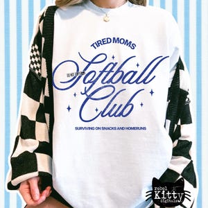 May include: White sweatshirt with the text "TIRED MOMS Softball Club" in blue script. The sweatshirt also has the phrase "SURVIVING ON SNACKS AND HOMERUNS" below the club name. The sleeves are partially covered by a black and white checkered cardigan.