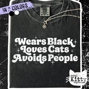 May include: Black t-shirt with the text "Wears Black Loves Cats Avoids People" in white. A gold necklace with a pendant is on the shirt. The shirt has a distressed look. The words "IN 2 COLORS" are in a purple banner.