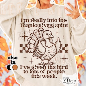 May include: A beige crewneck sweatshirt featuring a brown graphic of a turkey giving the middle finger. The text reads: I'm really into the Thanksgiving spirit. I've given the bird to lots of people this week. The design includes star and checkerboard accents.