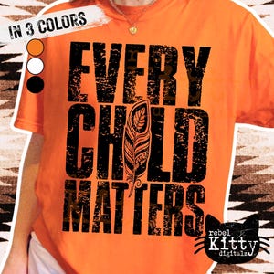May include: Orange t-shirt with the text "EVERY CHILD MATTERS" in large, distressed black letters. A detailed feather graphic is centered within the text. The shirt also features the text "IN 3 COLORS" and a logo of a black cat.