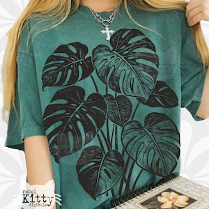 Op de afbeelding: Petrolgroen t-shirt met een grote zwarte illustratie van monstera bladeren. Het shirt heeft een relaxte pasvorm en korte mouwen. Het ontwerp is een botanisch thema, perfect voor plantenliefhebbers. Het shirt heeft de tekst "rebel Kitty digitalz".