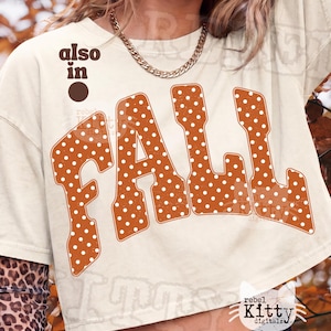 May include: A cream-colored short-sleeve t-shirt featuring the word FALL in large, orange, polka-dotted letters. Above the text, the words also in are printed in brown, accompanied by a brown circle. The shirt has a crew neckline.