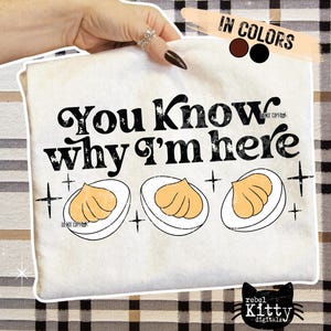 May include: Cream-colored t-shirt with the text "You Know why I'm here" in black. Below the text are three illustrations of deviled eggs. The shirt is held by a hand with a ring.