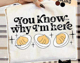 May include: Cream-colored t-shirt with the text "You Know why I'm here" in black. Below the text are three illustrations of deviled eggs. The shirt is held by a hand with a ring.