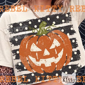 Peut inclure: Un t-shirt couleur crème arborant un grand motif de citrouille d'Halloween orange avec une tige verte. La citrouille est centrée sur une bande horizontale noire à pois blancs. Le texte REBEL KITTY est visible en orange en haut et en bas.