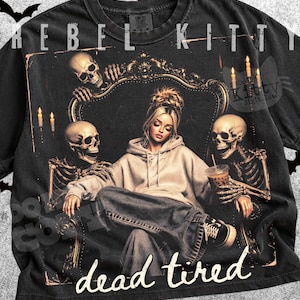May include: A black t-shirt featuring a graphic print of a person in a hoodie sitting on a throne flanked by two skeletons. The design includes lit candles, a coffee cup, and the text REBEL KITTY and dead tired in a white script font.