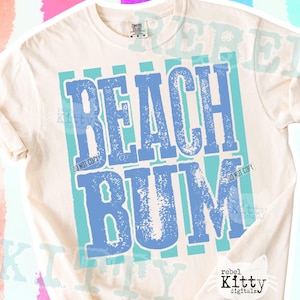 May include: Cream-colored t-shirt with the words "BEACH BUM" in distressed blue lettering. The text is set against a backdrop of vertical turquoise stripes, creating a summery vibe. The shirt is a Comfort Colors brand.