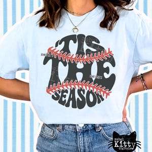 May include: Light blue t-shirt with a baseball graphic. The graphic features the words "TIS THE SEASON" in a baseball design with red stitching. The shirt is paired with blue denim jeans.