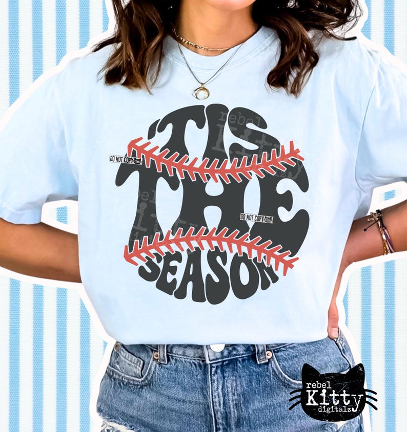 May include: Light blue t-shirt with a baseball graphic. The graphic features the words "TIS THE SEASON" in a baseball design with red stitching. The shirt is paired with blue denim jeans.