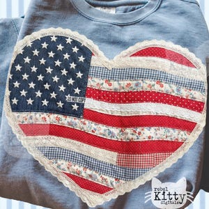 May include: A pale blue sweatshirt with a heart-shaped appliqué of the American flag. The flag design incorporates denim, floral patterns, and lace. The flag features white stars on a blue field and red and white stripes. The text "DO NOT COPY" is visible on the patch.