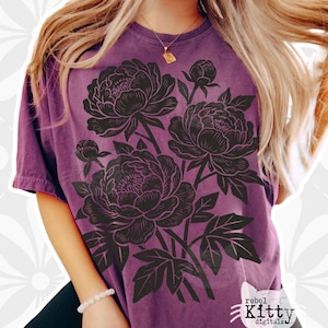 Puede incluir: Una camiseta morada con un diseño floral negro de peonías. El gráfico incluye tres grandes flores, capullos y hojas. La camiseta tiene cuello redondo y mangas cortas. El texto "rebel Kitty digitalz" está en la esquina inferior derecha.