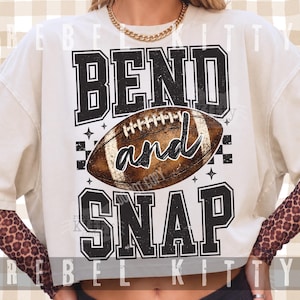 Op de afbeelding: Een crèmekleurig T-shirt met korte mouwen en een grafische print met de tekst BEND and SNAP in zwarte varsity-stijl letters. Een bruine American football staat centraal tussen de woorden, vergezeld door kleine ster- en dambordpatronen.