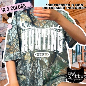 May include: Camouflage-patterned t-shirt with the word "HUNTING" in white block letters and "WIFE" below. The shirt has a distressed look and is available in three colors. The image also includes the text "DISTRESSED & NON DISTRESSED INCLUDED".