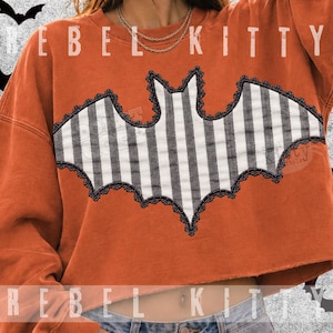 May include: An orange long-sleeve cropped sweatshirt featuring a large central bat applique. The bat has a black and white vertical striped pattern with a decorative black lace trim along its edges. The text REBEL KITTY is printed in white.