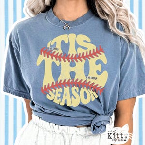 May include: A blue t-shirt with the text "'Tis the Season" in a baseball-themed design. The text is in yellow with a red baseball stitch design. The shirt has short sleeves and a crew neck.