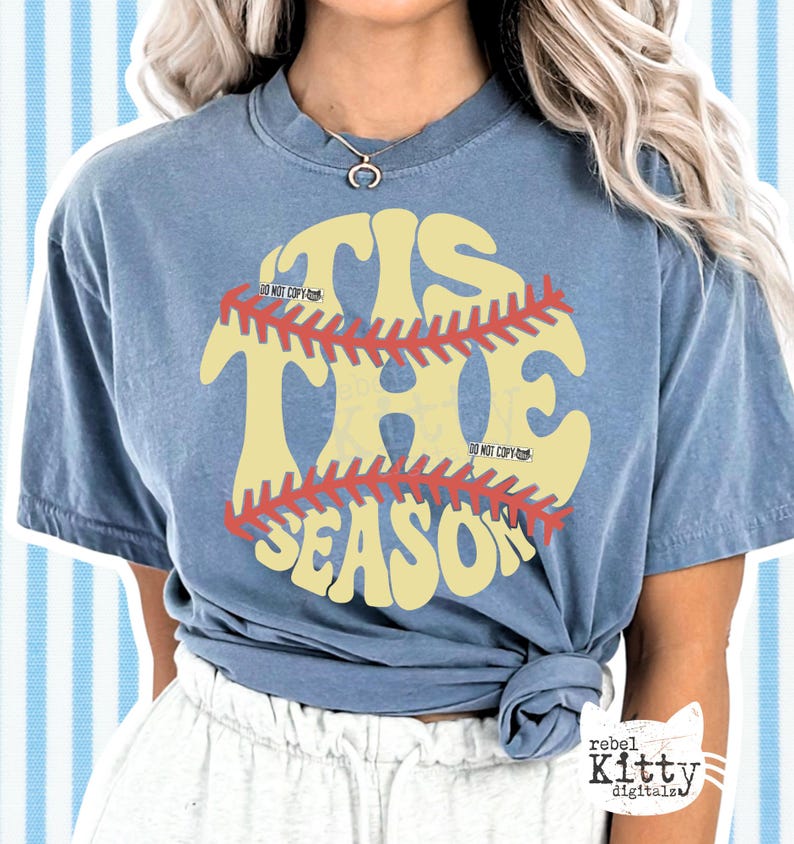 May include: A blue t-shirt with the text "'Tis the Season" in a baseball-themed design. The text is in yellow with a red baseball stitch design. The shirt has short sleeves and a crew neck.