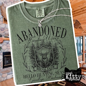 May include: Sage green t-shirt with the words "ABANDONED Hunting Wives Social Club" and a graphic of a deer within a shield. The shirt also has the phrases "Hello Hunting Seas Goodbye Husband". A silver necklace is draped across the shirt.