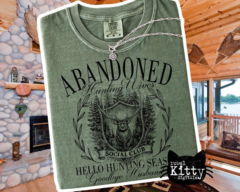 May include: Sage green t-shirt with the words "ABANDONED Hunting Wives Social Club" and a graphic of a deer within a shield. The shirt also has the phrases "Hello Hunting Seas Goodbye Husband". A silver necklace is draped across the shirt.