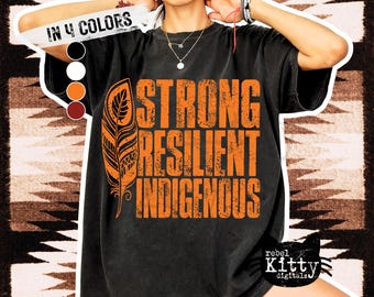 May include: Black t-shirt with the words "STRONG RESILIENT INDIGENOUS" in orange block letters and an orange feather graphic. The shirt also has the text "IN 4 COLORS" and the "rebel Kitty digitals" logo.