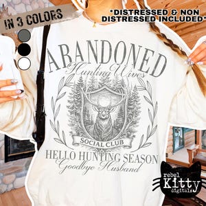 May include: Cream-colored sweatshirt with a graphic design featuring a deer, trees, and the words "ABANDONED Hunting Wives Social Club." The text "HELLO HUNTING SEASON Goodbye Husband" is also present. The image includes color options.