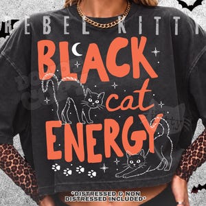 May include: A black short-sleeved t-shirt with a graphic print featuring the orange text "BLACK cat ENERGY" and two white line-art illustrations of cats. The design includes small white stars, a crescent moon, and paw prints. The shirt has a faded finish.