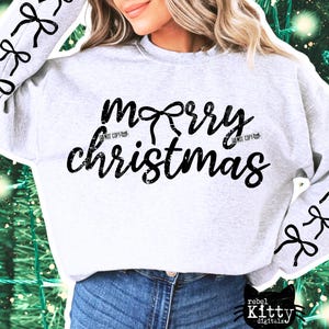 May include: A light gray sweatshirt with the words "merry christmas" in black script. The word "merry" has a bow design above it. Black bow designs are on the sleeves. The sweatshirt is paired with blue jeans.