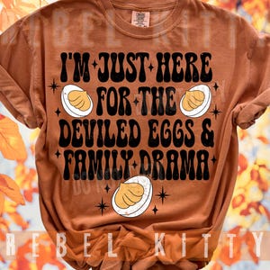 May include: A rust-colored short-sleeve t-shirt featuring black text that reads I'M JUST HERE FOR THE DEVILED EGGS & FAMILY DRAMA. The design includes three illustrations of halved deviled eggs and small black starburst accents across the front.