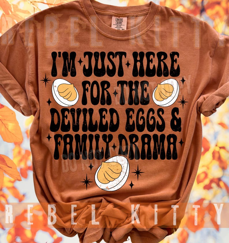 May include: A rust-colored short-sleeve t-shirt featuring black text that reads I'M JUST HERE FOR THE DEVILED EGGS & FAMILY DRAMA. The design includes three illustrations of halved deviled eggs and small black starburst accents across the front.