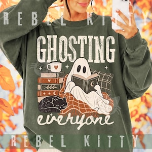 May include: A dark olive green crewneck sweatshirt featuring a graphic print with the text GHOSTING everyone. The design shows a white ghost reading a book, a stack of books, a coffee mug with a heart, and a black cat resting on a plaid blanket.