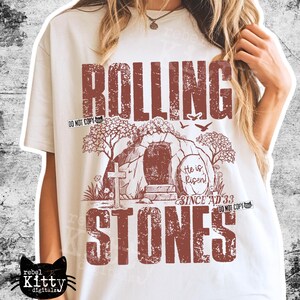 May include: Cream-colored t-shirt with a distressed brown graphic. The graphic features the words "ROLLING STONES" above and below an illustration of a tomb with a cross and the text "He is Risen! Since AD 33".