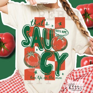 Op de afbeelding: Crèmekleurig T-shirt met een grafisch ontwerp. Het ontwerp toont de woorden "that's saucy" in groen en rood, samen met illustraties van tomaten. De zin "live life on the vine" is ook opgenomen. Het shirt heeft een relaxte pasvorm.