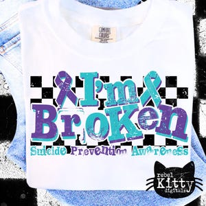 May include: White t-shirt with the words "I'm Broken" in teal and purple, with a checkered background and suicide prevention awareness text. Features a purple ribbon graphic.