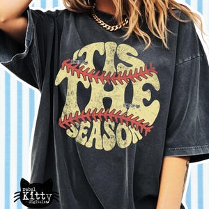 May include: Black t-shirt with a distressed graphic. The graphic features the words "TIS THE SEASON" in a vintage style, with a baseball design. The shirt has a relaxed fit and short sleeves. The Rebel Kitty Digitals logo is in the bottom left corner.